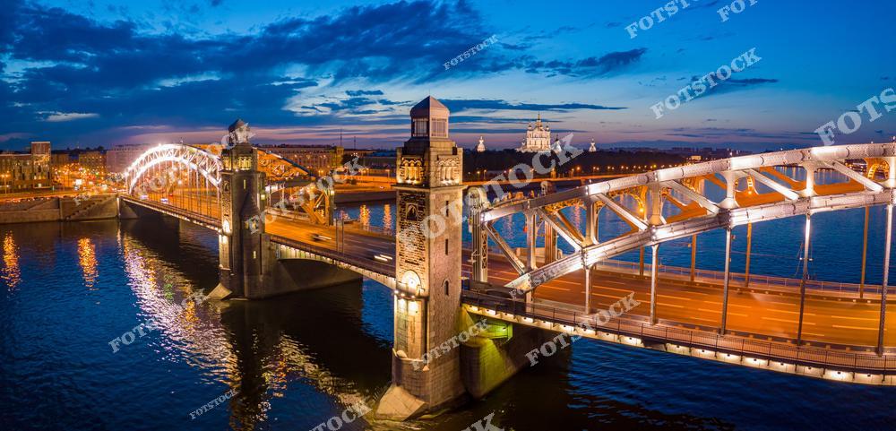 Панорамная фотография ночного Санкт-Петербурга с высоты птичьего полета. На снимке видны подсвеченные мосты города, включая Большеохтинский мост и мост Петра Великого, отражения огней в воде Невы и вечерние улицы. Съемка с дрона позволяет оценить архитектуру мостов, масштаб городской инфраструктуры и красоту вечернего Петербурга. Фотография передает атмосферу ночного города, праздничную иллюминацию и величие Северной столицы России. Снимок подходит для туристических публикаций, фотостоков, рекламы, оформления интерьеров и материалов о городских достопримечательностях.